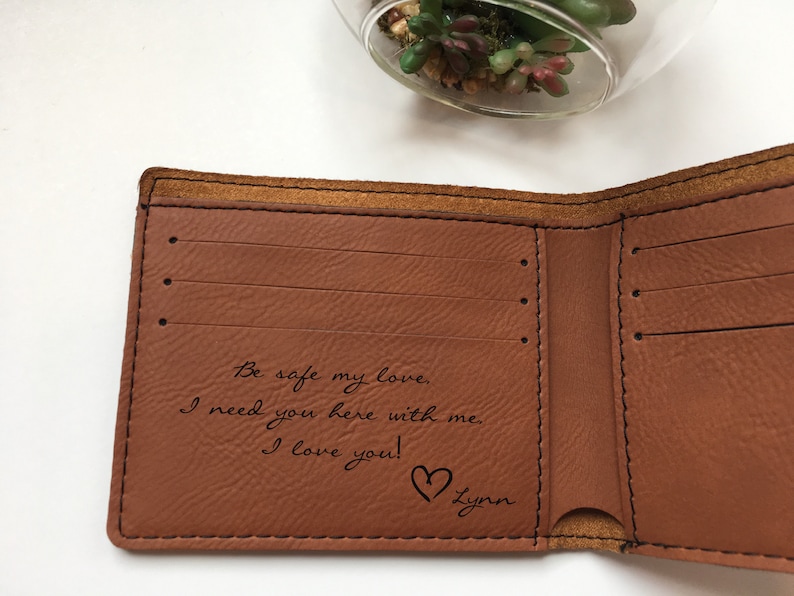 May include: A tan leather bifold wallet with black stitching and multiple card slots. The interior features a laser-engraved message that reads: Be safe my love. I need you here with me. I love you! followed by a heart symbol and the name Lynn.