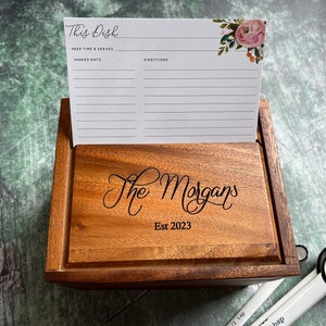 May include: A wooden recipe box with a lid engraved with the text The Morgans Est 2023. A white recipe card with floral accents and the text This Dish, Prep Time & Serves, Ingredients, and Directions is standing upright inside the box.