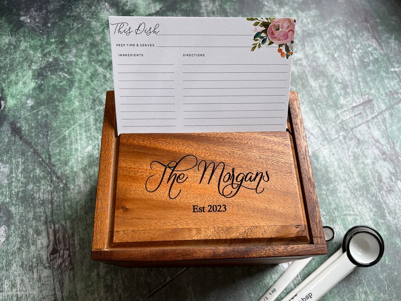 May include: A wooden recipe box with a lid engraved with the text The Morgans Est 2023. A white recipe card with floral accents and the text This Dish, Prep Time & Serves, Ingredients, and Directions is standing upright inside the box.