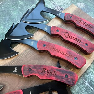May include: Four personalized black and red hatchet knives with wooden handles. The handles are engraved with the names "Ryan", "Solomonson", "Quinn", and "Molitor".