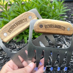 May include: Two stainless steel meat shredder claws with wooden handles. The handles are engraved with "Richie's Barbecue". The claws have blue protective tips. The tools are designed for shredding cooked meats.