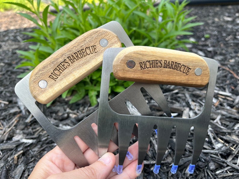 May include: Two stainless steel meat shredder claws with wooden handles. The handles are engraved with "Richie's Barbecue". The claws have blue protective tips. The tools are designed for shredding cooked meats.