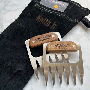 May include: Black grilling glove with "Keith Jr" text and two stainless steel meat shredder claws with wooden handles. One claw has "Life's a game. BBQ is serious." and the other has "SHRED IT BILLY" engraved.