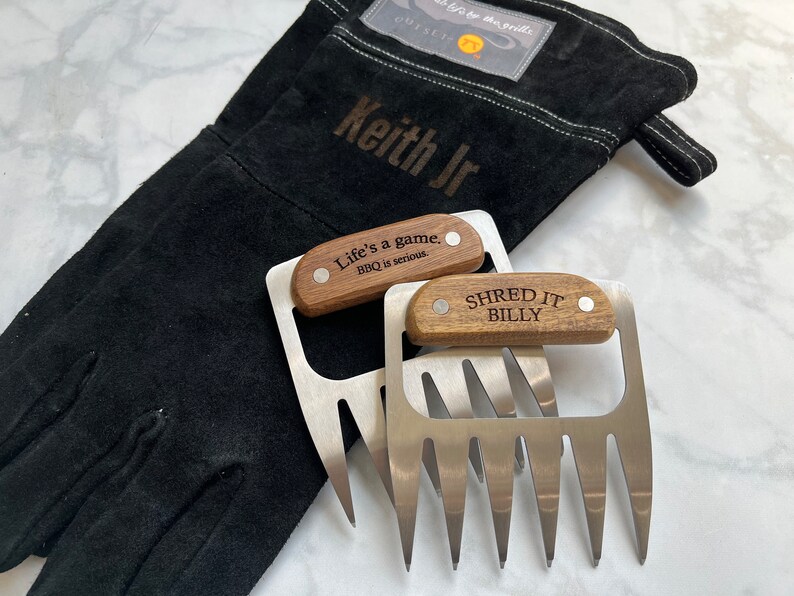 May include: Black grilling glove with "Keith Jr" text and two stainless steel meat shredder claws with wooden handles. One claw has "Life's a game. BBQ is serious." and the other has "SHRED IT BILLY" engraved.