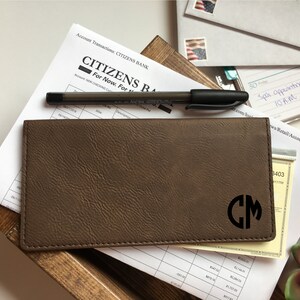 Puede incluir: Una funda rectangular marr&oacute;n para chequera con superficie texturizada y las letras negras del monograma CM grabadas en la esquina inferior derecha. La funda est&aacute; sobre un extracto de cuenta de Citizens Bank y un bol&iacute;grafo negro.