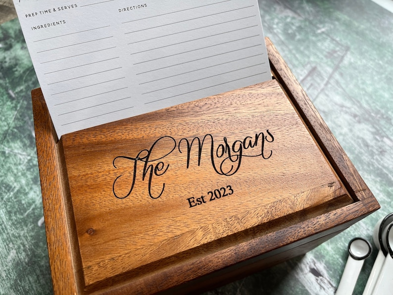 May include: A rectangular wooden recipe box with a dark brown finish, featuring the engraved text "The Morgans" and "Est 2023" on the front panel. A white recipe card with printed lines for ingredients and directions is tucked inside the box.