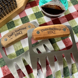 May include: Two stainless steel meat claws with wooden handles engraved with "Dad's Backyard BBQ" are displayed. A wooden grill brush and a small container of sauce are also visible on a red and green checkered cloth.