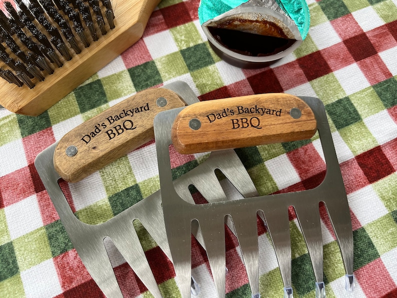 May include: Two stainless steel meat claws with wooden handles engraved with "Dad's Backyard BBQ" are displayed. A wooden grill brush and a small container of sauce are also visible on a red and green checkered cloth.