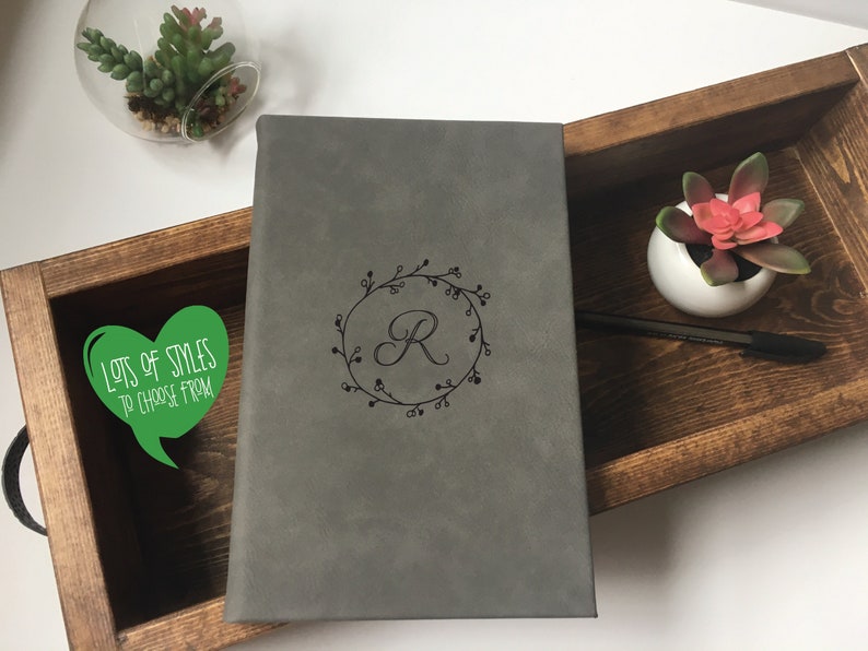 Puede incluir: Un diario de cuero gris con una corona de flores y la letra "R" grabada en la parte delantera. El diario est&aacute; en una bandeja de madera con una peque&ntilde;a planta suculenta en una maceta blanca.