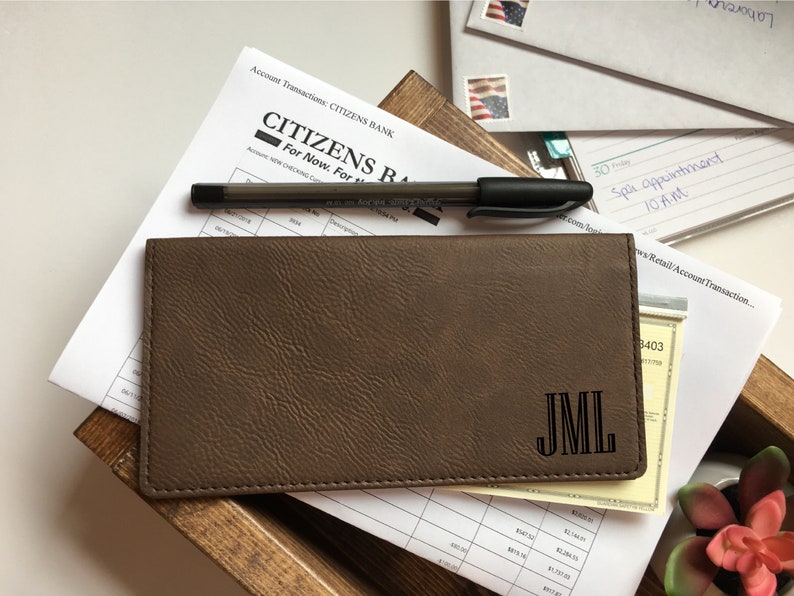 Puede incluir: Una funda rectangular para chequera de color marr&oacute;n con una superficie texturizada y el monograma JML grabado en la esquina inferior derecha. La funda est&aacute; colocada sobre un estado de cuenta de Citizens Bank y un bol&iacute;grafo negro, con costuras visibles.