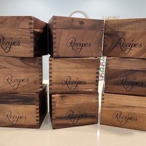 May include: Nine wooden recipe boxes stacked in three rows of three. Each box is a dark brown color and has the word "Recipes" engraved on the front in a cursive font.