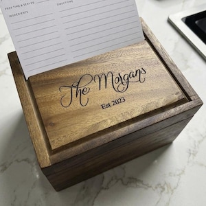 May include: A rectangular wooden recipe box with a hinged lid featuring the engraved text The Morgans Est 2023. A white recipe card with printed fields for prep time, serves, ingredients, and directions is inserted into the lid slot.