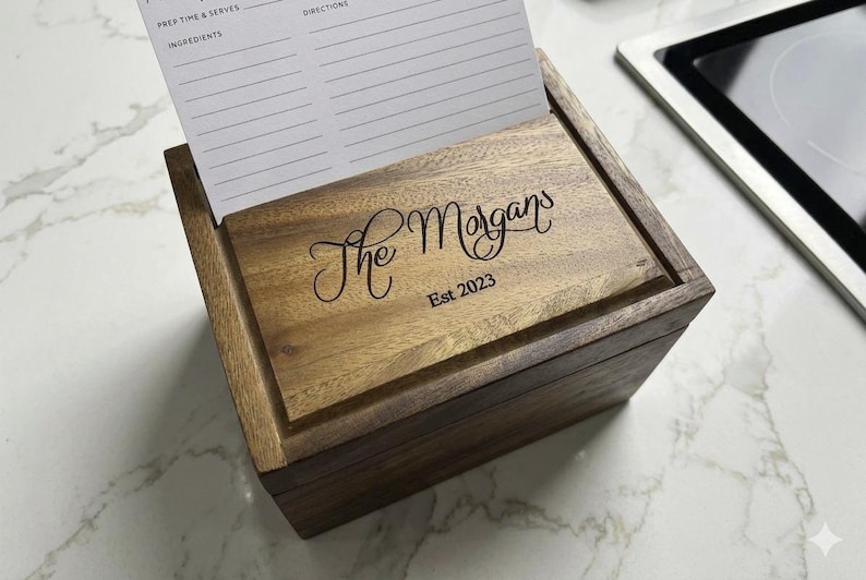 May include: A rectangular wooden recipe box with a hinged lid featuring the engraved text The Morgans Est 2023. A white recipe card with printed fields for prep time, serves, ingredients, and directions is inserted into the lid slot.