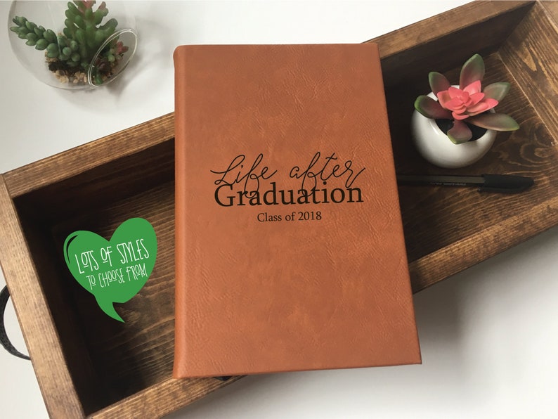 Puede incluir: Un diario de cuero marr&oacute;n con el texto "Life after Graduation Class of 2018" grabado en la portada. El diario est&aacute; en una caja de madera con una pegatina verde en forma de coraz&oacute;n que dice "Lots of Styles to Choose From".