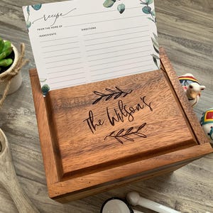 May include: A wooden recipe box with a hinged lid engraved with the text The Wilson's and leaf motifs. A white recipe card with eucalyptus illustrations is tucked inside. The box is made of brown wood with a visible grain and a smooth finish.