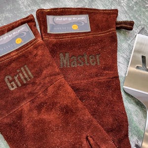 K&ouml;nnte beinhalten: Zwei braune Grillhandschuhe aus Leder mit den gepr&auml;gten W&ouml;rtern "Grill" und "Master". Ein Edelstahl-Pfannenwender liegt neben den Handschuhen. Jeder Handschuh hat ein Etikett mit der Aufschrift "Grab life by the grills".
