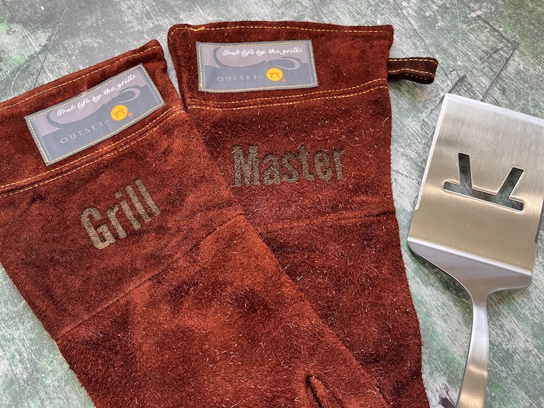 K&ouml;nnte beinhalten: Zwei braune Grillhandschuhe aus Leder mit den gepr&auml;gten W&ouml;rtern "Grill" und "Master". Ein Edelstahl-Pfannenwender liegt neben den Handschuhen. Jeder Handschuh hat ein Etikett mit der Aufschrift "Grab life by the grills".