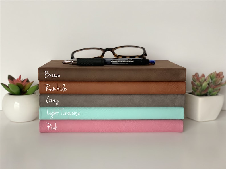 Puede incluir: Una pila de cinco diarios encuadernados en cuero de diferentes colores: marr&oacute;n, crudo, gris, turquesa claro y rosa. Los diarios est&aacute;n apilados sobre una superficie blanca con un bol&iacute;grafo y unas gafas en la parte superior.
