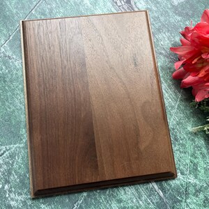 May include: A rectangular, dark brown wooden plaque with a beveled edge. The wood grain is visible, and the plaque is set against a green and white marbled surface. A red flower with green foliage is in the upper right corner.
