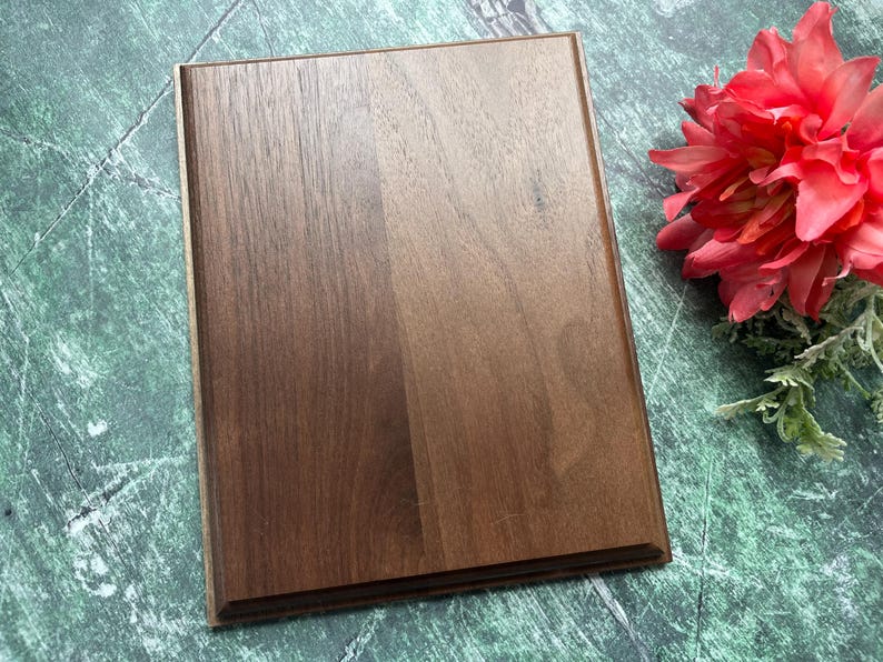 May include: A rectangular, dark brown wooden plaque with a beveled edge. The wood grain is visible, and the plaque is set against a green and white marbled surface. A red flower with green foliage is in the upper right corner.