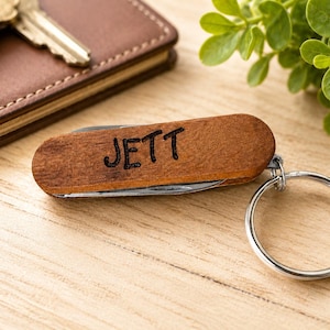 May include: A brown wooden pocket knife keychain with the name JETT engraved in black lettering on the side. The multi-tool features a silver metal keyring attached to one end, with visible metal components tucked inside the wooden handle casing.