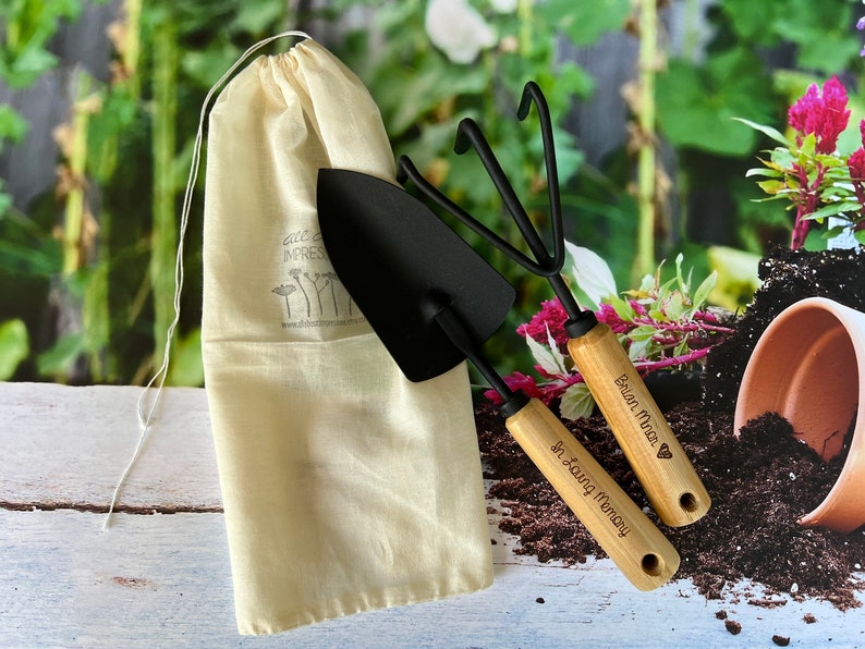 May include: A set of two gardening tools, a trowel and a hand rake, with wooden handles engraved with the text "In Loving Memory" and "Brian Finch" with a heart symbol. The tools are in a white drawstring bag.