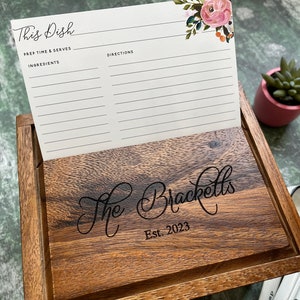 May include: A dark wood recipe box with a hinged lid engraved with the text The Bracketts Est. 2023. A white recipe card with floral accents and the text This Dish, Prep Time & Serves, Ingredients, and Directions is displayed in the open lid slot.