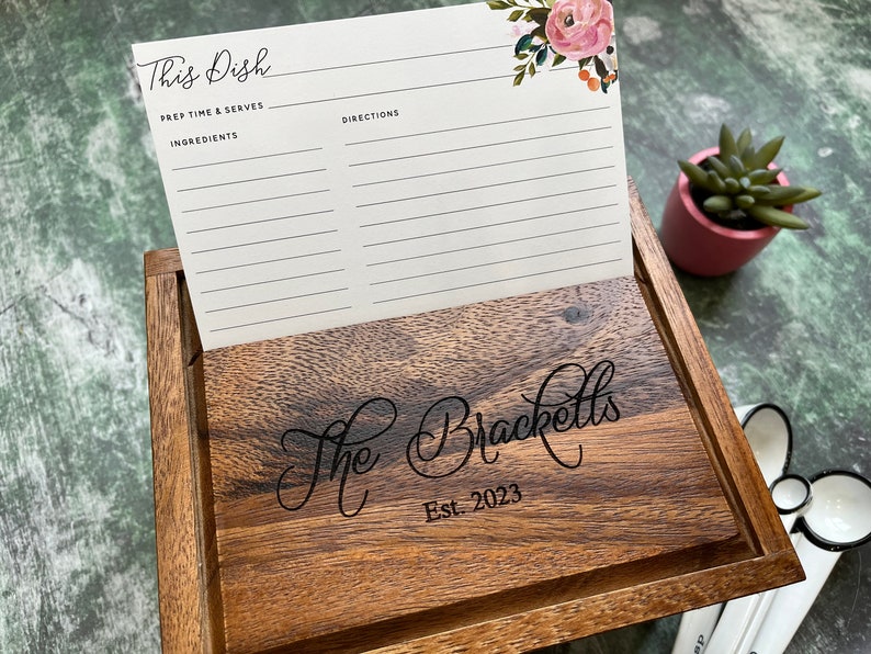 May include: A dark wood recipe box with a hinged lid engraved with the text The Bracketts Est. 2023. A white recipe card with floral accents and the text This Dish, Prep Time & Serves, Ingredients, and Directions is displayed in the open lid slot.