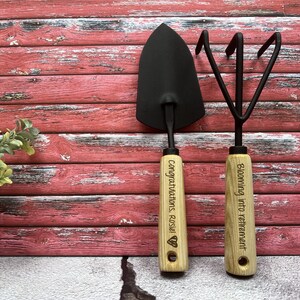 May include: A set of two gardening tools with wooden handles. The handle of the trowel is engraved with "Congratulations Rosie!" and the handle of the cultivator is engraved with "Blooming into retirement".