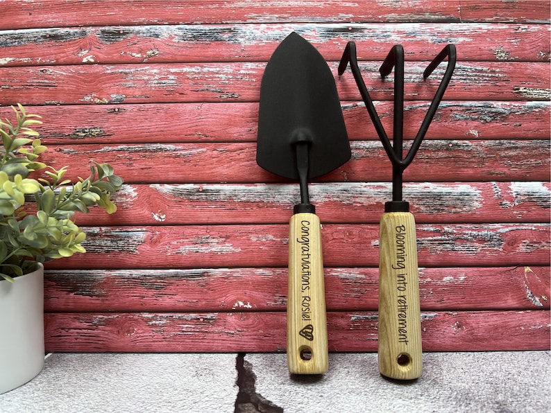 May include: A set of two gardening tools with wooden handles. The handle of the trowel is engraved with "Congratulations Rosie!" and the handle of the cultivator is engraved with "Blooming into retirement".