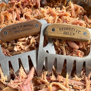 May include: Two stainless steel meat shredder claws with wooden handles rest on a bed of pulled pork. One handle reads "Life's a game. BBQ is serious." The other reads "SHRED IT BILLY."