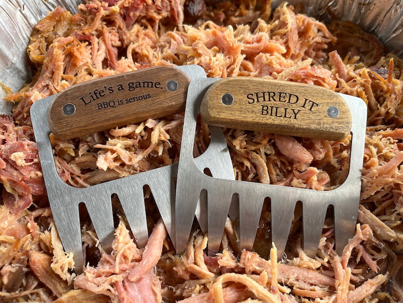 May include: Two stainless steel meat shredder claws with wooden handles rest on a bed of pulled pork. One handle reads "Life's a game. BBQ is serious." The other reads "SHRED IT BILLY."