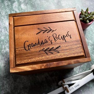 May include: A rectangular wooden box with a hinged lid featuring the engraved text Grandma's Recipes surrounded by two leaf sprigs. The box has a natural brown wood grain finish and is accompanied by a set of white plastic measuring spoons.