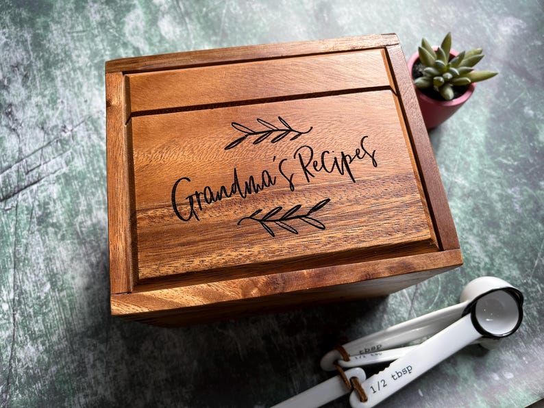 May include: A rectangular wooden box with a hinged lid featuring the engraved text Grandma's Recipes surrounded by two leaf sprigs. The box has a natural brown wood grain finish and is accompanied by a set of white plastic measuring spoons.