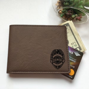 May include: A brown bi-fold wallet with a textured surface featuring a black engraved police badge emblem on the front. The wallet contains a United States twenty-dollar bill and a Bank of America credit card tucked inside the open side pocket.