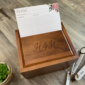 Pu&ograve; includere: Una scatola di legno per ricette con un design floreale sul coperchio. La scatola &egrave; incisa con le iniziali "H & H". La scatola &egrave; aperta e all'interno &egrave; visibile un cartoncino per ricette vuoto. Ci sono cucchiai dosatori sul tavolo accanto alla scatola.