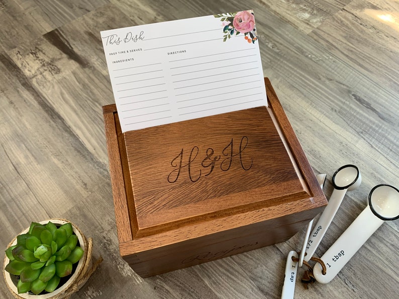 Pu&ograve; includere: Una scatola di legno per ricette con un design floreale sul coperchio. La scatola &egrave; incisa con le iniziali "H & H". La scatola &egrave; aperta e all'interno &egrave; visibile un cartoncino per ricette vuoto. Ci sono cucchiai dosatori sul tavolo accanto alla scatola.
