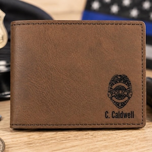 May include: A brown bi-fold wallet featuring a dark brown engraved police badge emblem and the text C. Caldwell printed in black. The wallet has visible dark brown stitching along the edges and a smooth, matte-textured surface finish.
