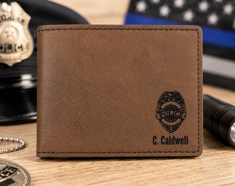 May include: A brown bi-fold wallet featuring a dark brown engraved police badge emblem and the text C. Caldwell printed in black. The wallet has visible dark brown stitching along the edges and a smooth, matte-textured surface finish.