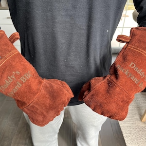 K&ouml;nnte beinhalten: Ein Paar braune Leder-BBQ-Handschuhe. Auf den Handschuhen steht der Text "Daddy's Backyard BBQ". Jeder Handschuh hat eine kleine Schlaufe zum Aufh&auml;ngen und einen Aufn&auml;her mit Text und Logo.