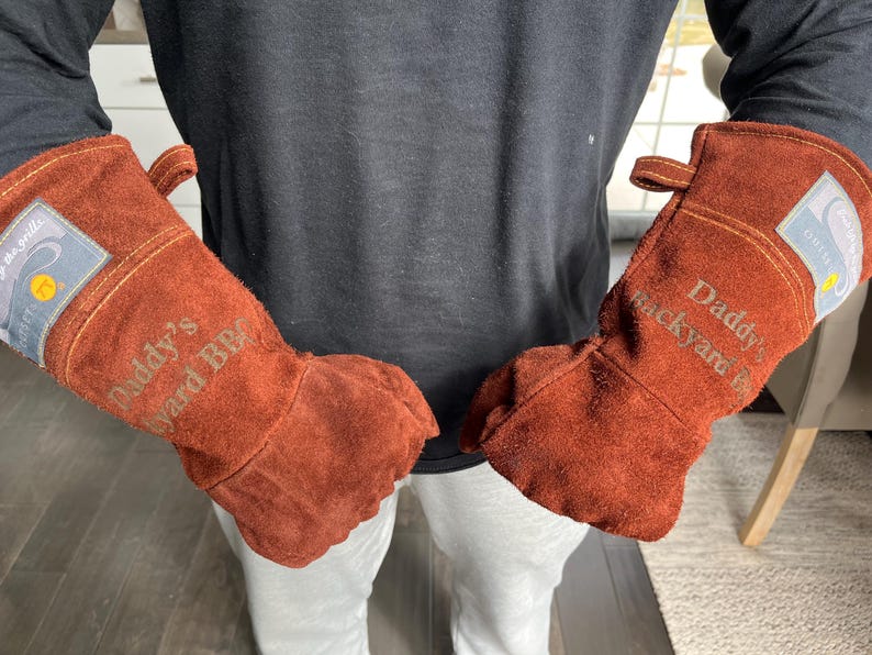 K&ouml;nnte beinhalten: Ein Paar braune Leder-BBQ-Handschuhe. Auf den Handschuhen steht der Text "Daddy's Backyard BBQ". Jeder Handschuh hat eine kleine Schlaufe zum Aufh&auml;ngen und einen Aufn&auml;her mit Text und Logo.