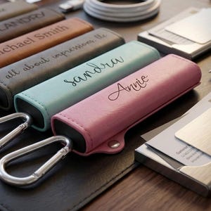 May include: A collection of personalized leather cases in various colors, each with a name or phrase engraved on them. The cases are displayed with carabiner clips, business card holders, and a pen on a dark surface.