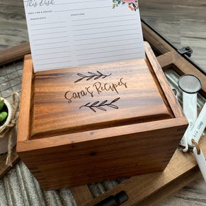 May include: A rectangular wooden recipe box with a lid engraved with the text "Sara's Recipes" and leaf motifs. A white recipe card with floral accents is tucked into the lid. Beside the box are white measuring spoons with black rims.