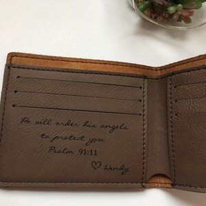 May include: A brown bi-fold wallet with a smooth interior featuring three horizontal card slots on the left side. The interior is engraved with the text: He will order his angels to protect you. Psalm 91:11, followed by a heart icon and the name Wendy.