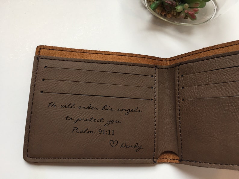 May include: A brown bi-fold wallet with a smooth interior featuring three horizontal card slots on the left side. The interior is engraved with the text: He will order his angels to protect you. Psalm 91:11, followed by a heart icon and the name Wendy.