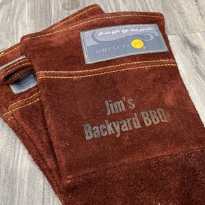 A pair of brown leather gloves with a label that says "Jim's Backyard BBQ" printed on them.