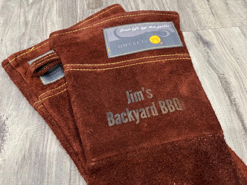 A pair of brown leather gloves with a label that says "Jim's Backyard BBQ" printed on them.