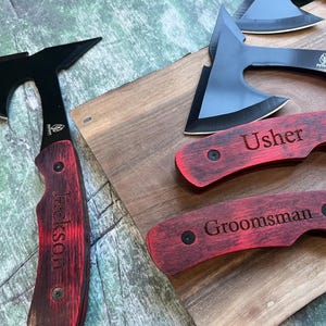 May include: A set of three personalized pocket knives with black blades and red wooden handles. The handles are engraved with the words "Usher", "Groomsman", and "Jackson".