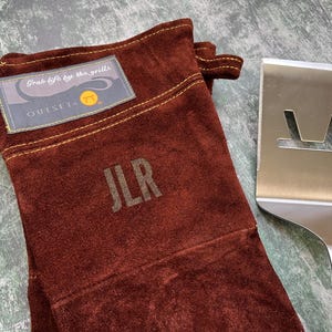 K&ouml;nnte beinhalten: Ein brauner Grillhandschuh aus Wildleder mit den Initialen "JLR" und dem Text "Grab life by the grills" auf einem Aufn&auml;her. Ein Edelstahl-Pfannenwender mit einem Ausschnitt-Design befindet sich neben dem Handschuh. Der Handschuh hat eine Schlaufe zum Aufh&auml;ngen.