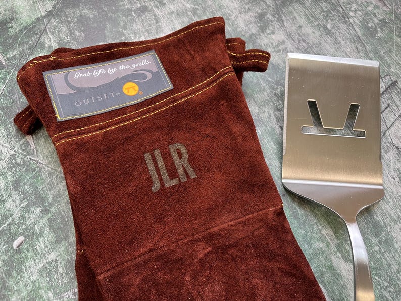 K&ouml;nnte beinhalten: Ein brauner Grillhandschuh aus Wildleder mit den Initialen "JLR" und dem Text "Grab life by the grills" auf einem Aufn&auml;her. Ein Edelstahl-Pfannenwender mit einem Ausschnitt-Design befindet sich neben dem Handschuh. Der Handschuh hat eine Schlaufe zum Aufh&auml;ngen.